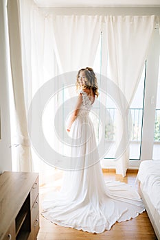 A sophisticated bride in an elegant wedding dress stands in the hotel room during preparation to the wedding ceremony