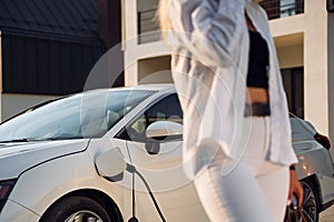With sopping bag. Young woman in white clothes is with her electric car at daytime