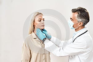 Sore throat. Professional mature doctor in medical protective mask checking thyroid glands of young beautiful woman