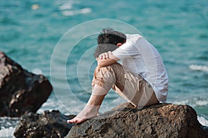 Sorrowful stressed Asian man sit and hug knees up to the chest at the rock of seashore.