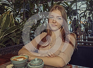 Sorrowful young lady is sitting in cafe, coffee cup is served on table in front of her