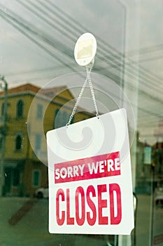 Sorry we are closed sign in red, hanging inside on shop glass door with city reflection.