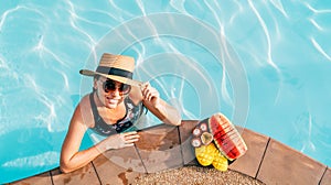 Soulful smiling woman in straw hat in sunglasses swimming in pool and enjoying fresh tropical fruits