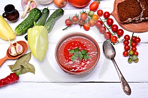 Soup gazpacho in a brown ceramic plate on a white wooden background, top view, around fresh vegetables and herbs for cooking