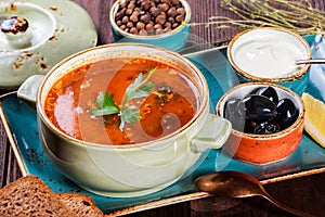 Soup with meat, olives, herbs, lemon, sour cream in bowl, black bread and spices on dark wooden background, homemade food.