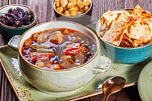 Soup with meat, oregano, chickpeas, peppers and vegetables served with crackers and bread on plate on dark wooden background.