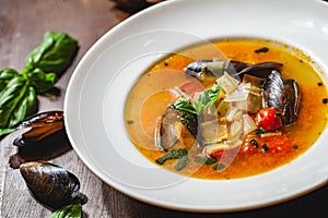 Soup with mussels and vegetables in white plate on wooden table.