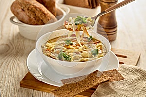 Soup with noodle, pieces of chicken and vegetables in white bowl served with spices and breads on light wooden background