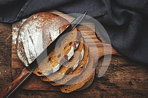 Sourdough rye bread with raisins, pieces on wooden cutting board, bread knife and black linen kitchen towel. Rustic table