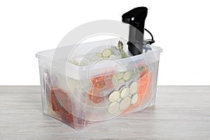 Sous vide cooker and vacuum packed food products in box on wooden table against white background. Thermal immersion circulator