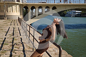 South American woman, young and beautiful, brunette, with brown dress, leaning on a railing with a relaxed and flirtatious