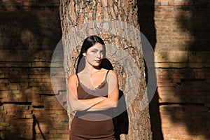 South American woman, young and beautiful, brunette, in brown dress, leaning on the trunk of a tree with her arms crossed, angry.