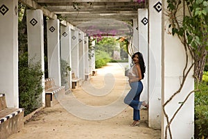 South American woman, young, beautiful, brunette, with crochet top and jeans leaning on a column, posing flirtatious and happy.