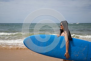 South American woman, young and beautiful, brunette with sunglasses and swimsuit, coming out of the water holding a blue surfboard