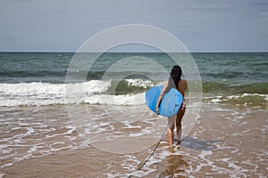 South American woman, young and beautiful, brunette with sunglasses and swimsuit, running into the water holding a blue surfboard