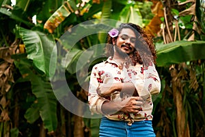 South Asian gay man with floral shirt, blue jeans stands in tropical foliage, purple flower in hair. Confident smile