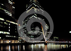 The South Quay Footbridge At South Dock In Front Of The Brightly Lit Bank Towers Of Canary Wharf London England At Night