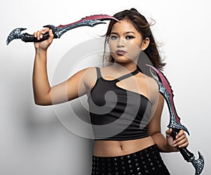 Southeast Asian girl in a black top and skirt with two fantasy knives against a white wall