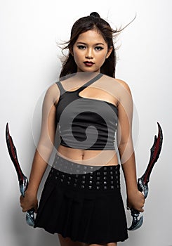 Southeast Asian girl in a black top and skirt with two fantasy knives against a white wall