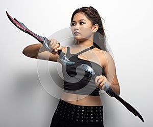 Southeast Asian girl in a black top and skirt with two fantasy knives against a white wall