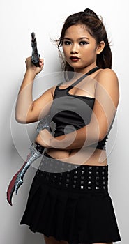 Southeast Asian girl in a black top and skirt with two fantasy knives against a white wall