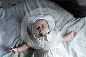 Southeast Asian newborn is laying on the white bed. South East Asian new born is wearing white hat and dress.
