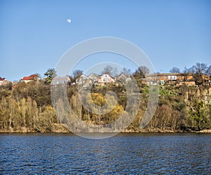 Southern Bug River in spring. On the cliff are houses, and in the blue sky the moon is visible