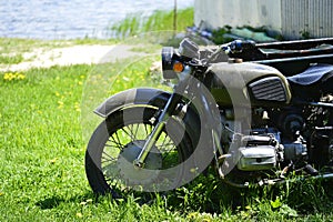 The Soviet Dnepr motorcycle on the green grass of the front part close up against a sandy shore by the lake