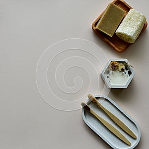 Soy candle, bamboo toothbrushes on a concrete tray, natural soap and loofah sponge bath on a wooden plate. Beige paper background.