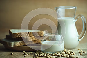 Soybean milk in a glass jug and whole wheat bread in wood disk on table.