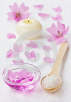 Spa composition with sea salt bath in wooden spoon, pink flowers petals and burning candle on a white surface