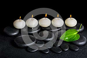 spa concept of row white candles and green leaf on black zen stones background with dew, closeup