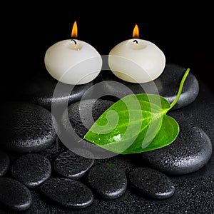 spa concept of two white candles and green leaf on black zen stones background with dew, closeup