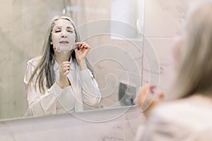 Spa at home, antiwrinkle facial mask. Pretty senior woman with long gray hair. standing in front of mirror in bathroom