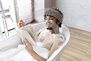 Spa procedure, self care and resting in bathroom. Happy young indian man in bath with foam, reading book in morning