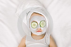 Spa procedures for a little cute baby girl in white bath towel with cucumbers on her eyes lying on white background