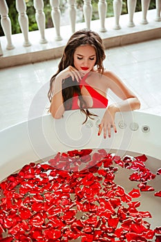 Spa Relax.  Beautiful Bikini Woman enjoying bath with red rose petals. Health And Beauty. Sexy Girl in red swimwear Bathing With