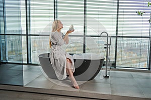 Spa relaxation. Young woman in spa bathrobe sitting at bath blowing foam in bathtub. Girl relaxing in bathroom at home