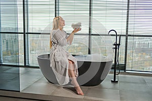 Spa relaxation. Young woman in spa bathrobe sitting at bath blowing foam in bathtub. Girl relaxing in bathroom at home