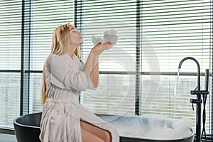 Spa relaxation. Young woman in spa bathrobe sitting at bath blowing foam in bathtub. Girl relaxing in bathroom at home