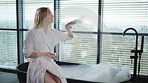 Spa relaxation. Young woman in spa bathrobe sitting at bath blowing foam in bathtub. Girl relaxing in bathroom at home