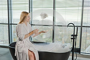 Spa relaxation. Young woman in spa bathrobe sitting at bath blowing foam in bathtub. Girl relaxing in bathroom at home