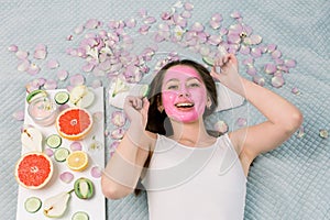 Spa, skin care, cosmetology concept. Pretty smiling woman with pink clay facial mask and cucumber slices in her hands
