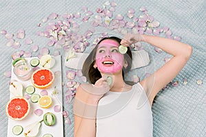 Spa, skin care, cosmetology concept. Pretty smiling woman with pink clay facial mask and cucumber slices in her hands