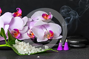 spa still life of sea salt on leaf, lilac orchid with drops and burning aroma incense cones over black zen stones