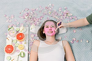 Spa therapy for young smiling woman receiving facial mask at beauty salon - indoors. Pretty girl with pink facial mask