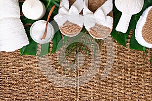 Spa tools: white towel, bamboo slippers, herbal ball, cream, wooden brush, coconut oil, monstera on rattan background. Cosmetic