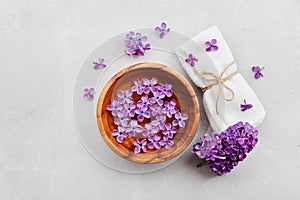 Spa and wellness composition with perfumed lilac flowers water in wooden bowl and towel on stone background, top view, flat lay