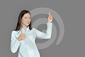 Space for Text or Product. Young woman standing isolated on grey pointing up thumb up smiling positive