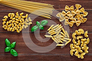 Spaghetti, farfalle, pipe rigatoni, maccheroni, penne rigate, cresta di gallo, raw, leaf of basil, on wooden brown planks dark photo
